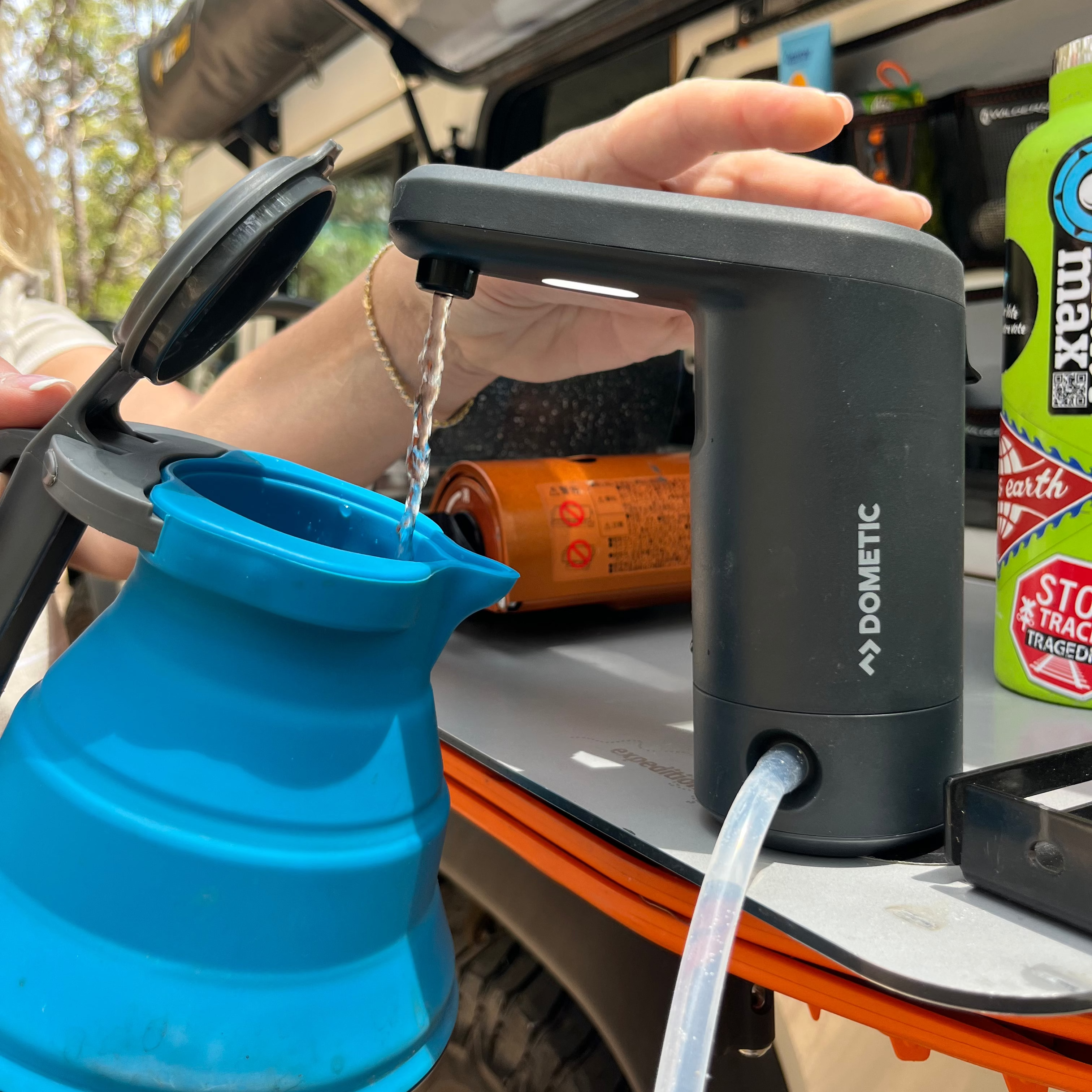 Camping water pump being used to fill a blue collapsible cup with a vehicle in the background.