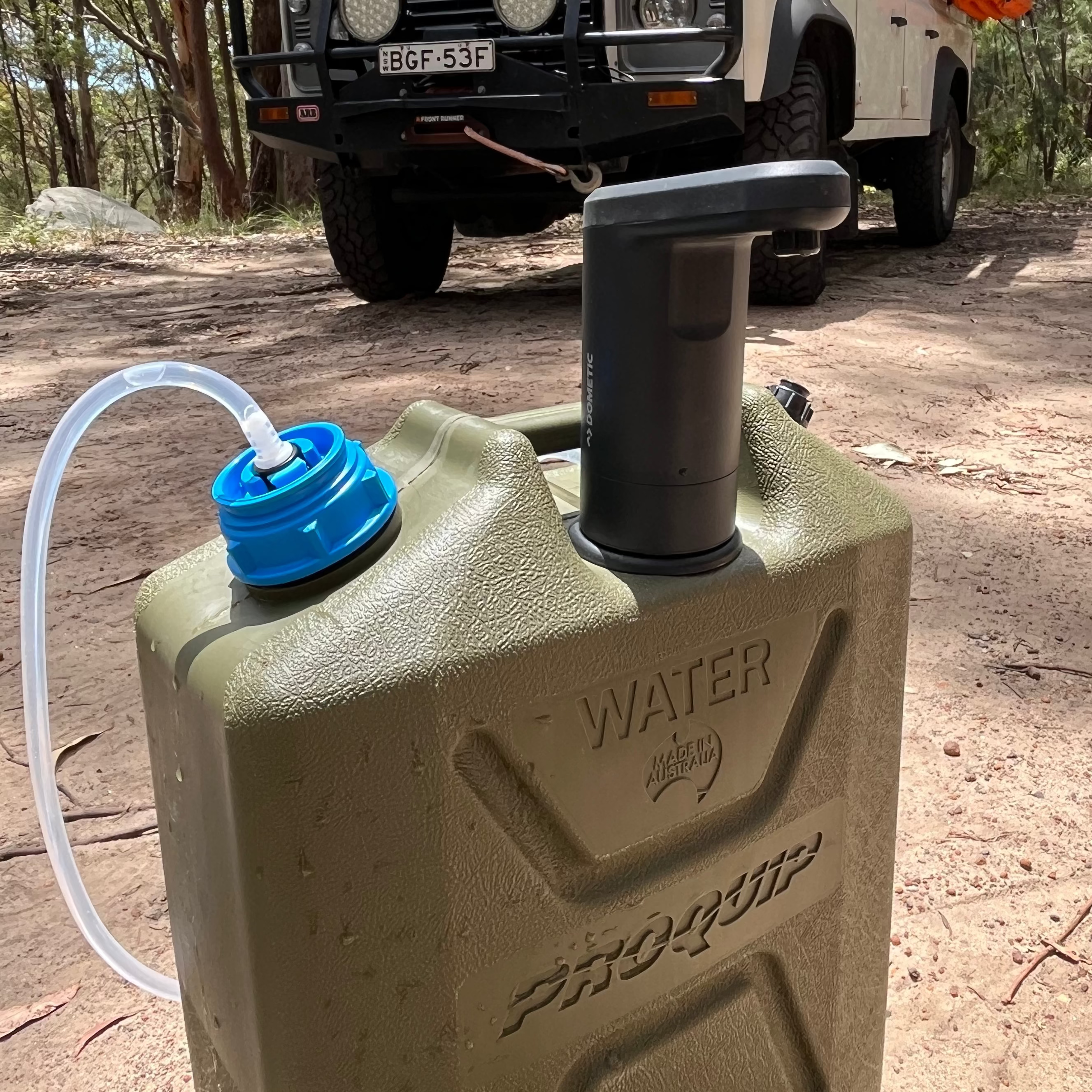 Green water container with a blue cap on a dirt road with a vehicle in the background.