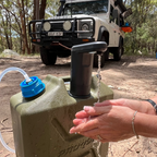Person washing hands with water from a green container outdoors, with a vehicle in the background.